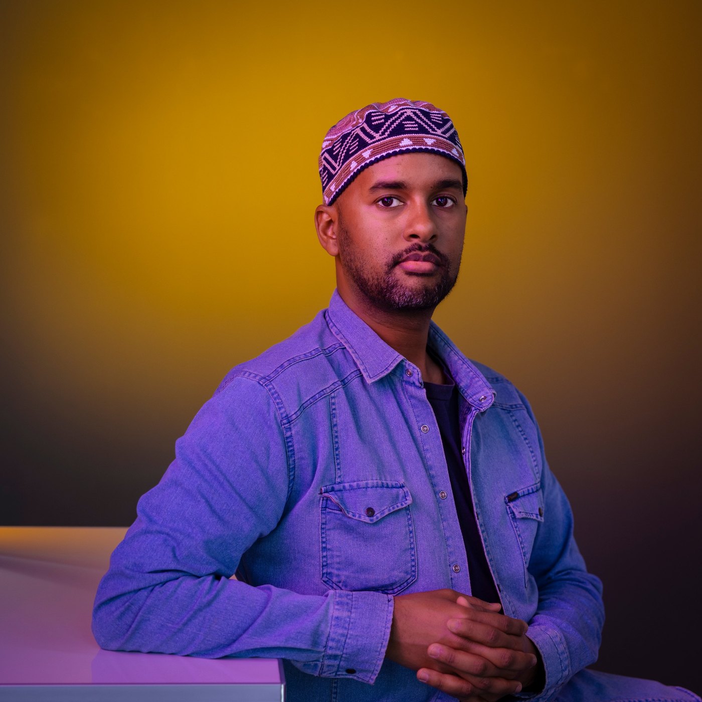 Portrait of Mustafa Saeed in a denim shirt and a patterned cap, against a yellow background